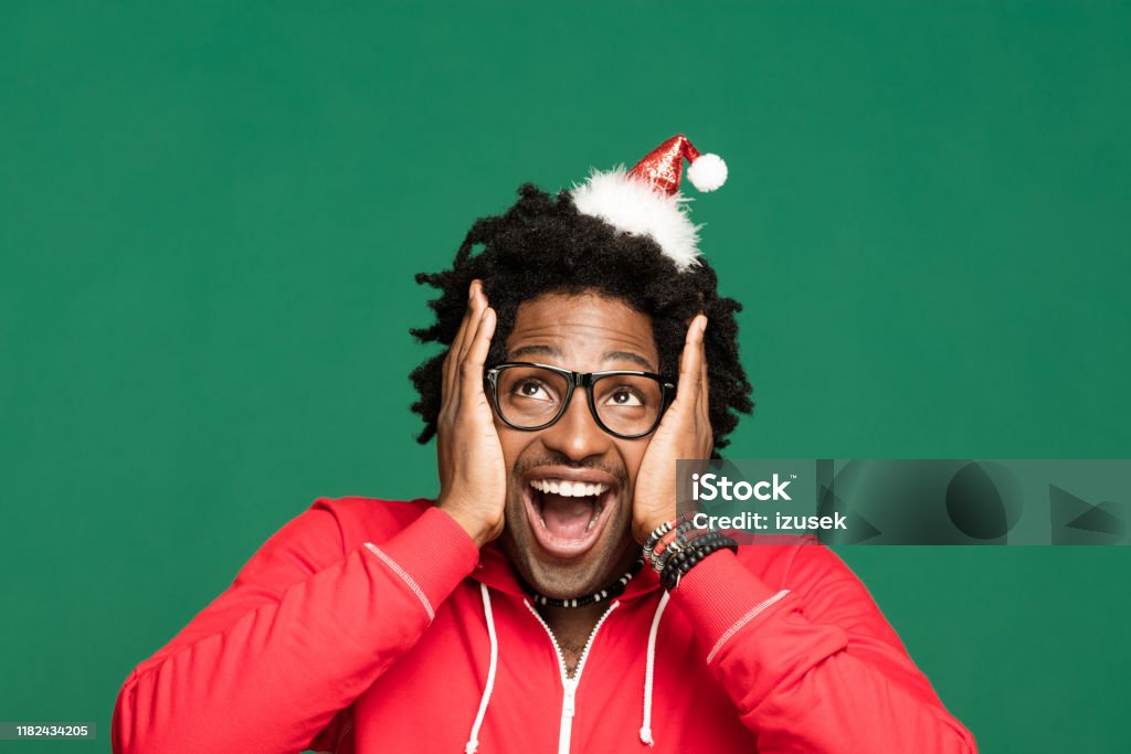Funny christmas portrait of excited young man wearing in red Funny Christmas portrait of young afro American man wearing Santa Claus hat headband and red blouse, looking away and laughing. Studio shot against green background. Christmas Stock Photo