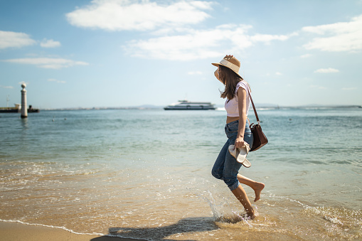 Barefoot young woman walking on the  beach in Lisbon at summer vacation, wearing sun hat