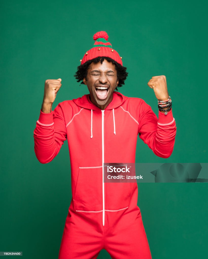 Funny christmas portrait of excited young man wearing in red pajamas Funny Christmas portrait of young afro American man wearing red pajamas and woolen hat, laughing with raised hands. Studio shot against green background. Christmas Stock Photo