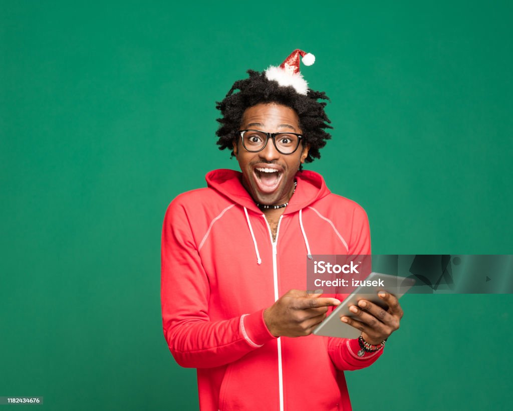Funny christmas portrait of excited young man wearing in red, holding digital tablet Funny Christmas portrait of young afro American man wearing Santa Claus hat headband and red onesies, holding digital tablet and looking at camera. Studio shot against green background. 25-29 Years Stock Photo