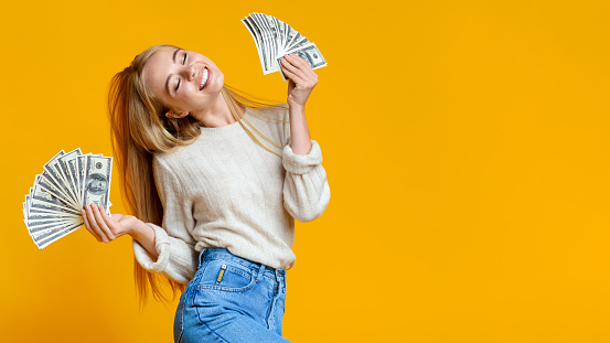Blissful millennial girl holding lots of money and enjoying success on orange studio background, panorama with free space