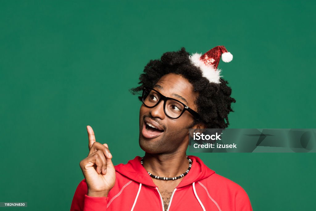 Funny christmas portrait of happy young man wearing in red Funny Christmas portrait of young afro American man wearing Santa Claus hat headband and red blouse, looking away and pointing at copy space. Studio shot against green background. Smiling Stock Photo