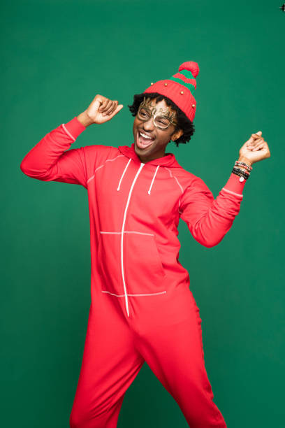 Funny christmas portrait of happy young man wearing in red pajamas Funny Christmas portrait of young afro American man wearing red pajamas and woolen hat, dancing and singing with raised hands. Studio shot against green background. babygro stock pictures, royalty-free photos & images