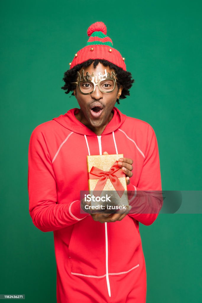 Funny christmas portrait of surprised young man holding present Funny Christmas portrait of young afro American man wearing red pajamas and woolen hat, holding gift in hands and laughing at camera. Studio shot against green background. Gift Stock Photo
