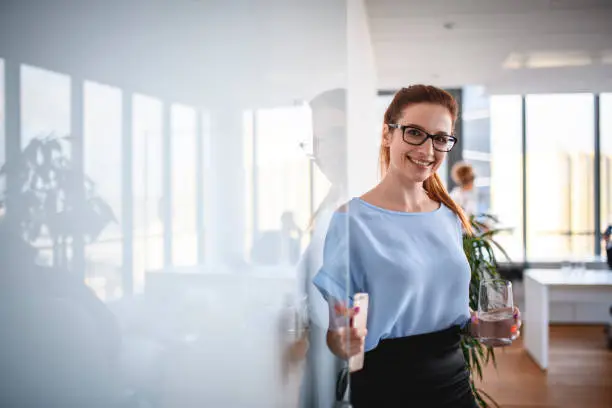 Bespectacled Caucasian businesswoman in mid 20s holding open front door to modern corporate office and looking at camera.