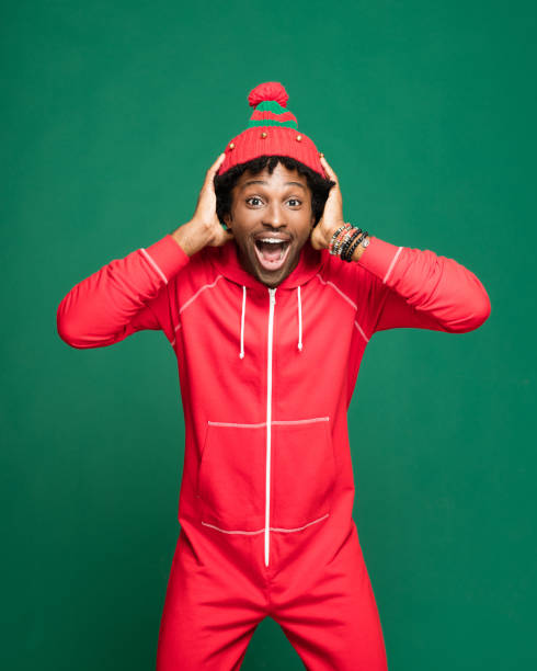 Funny christmas portrait of surprised young man wearing in red pajamas Funny Christmas portrait of young afro American man wearing red pajamas and woolen hat, laughing at camera with head in hands. Studio shot against green background. babygro stock pictures, royalty-free photos & images