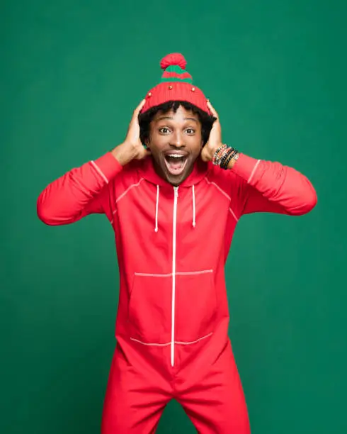 Funny Christmas portrait of young afro American man wearing red pajamas and woolen hat, laughing at camera with head in hands. Studio shot against green background.