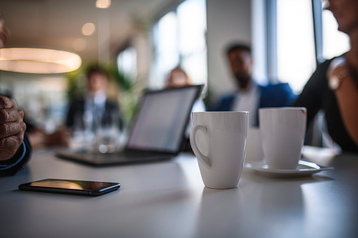 Foreground close-up of coffee mugs, smart phone, and laptop sitting on conference table between executive team members.