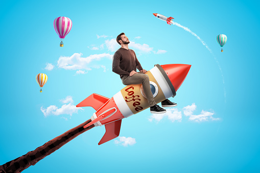 Confident young man riding toy rocket made of coffee cup in blue sky with hot air balloons and rocket in background. Food and drink advertising. Diet and nutrition. Face the day.