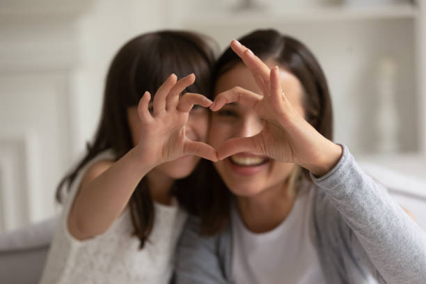 Happy young mother with little daughter making focused heart sign. Happy young mother with cute little daughter making focused heart sign with hands, looking at camera. Smiling millennial mom and small girl showing love gesture together, expressing care, affection. attached stock pictures, royalty-free photos & images