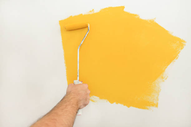 Construction worker and handyman doing finish renovation at apartment. Professional painter using paint roller brush painting of wall with yellow color on construction site. Home renovation concept.