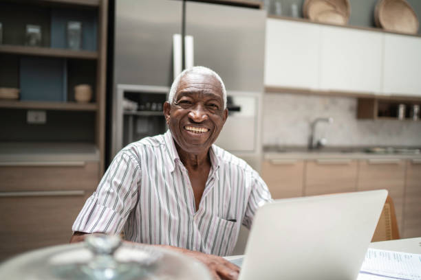 retrato de um homem sênior que usa o portátil na tabela de cozinha - laptop senior adult computer men - fotografias e filmes do acervo