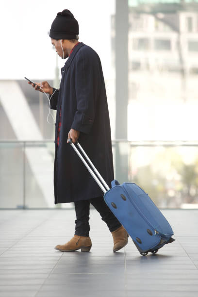 côté de corps complet de l'homme de voyage américain africain marchant avec le téléphone portable et la valise à la station - pulling a name from a hat photos et images de collection