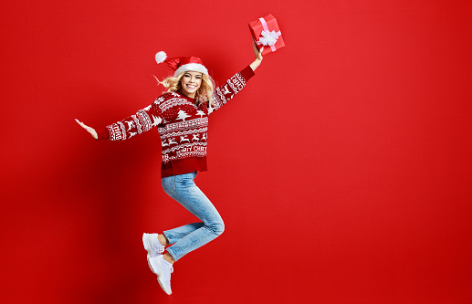 happy young cheerful girl laughs and jumps in christmas hat and with  gift on  red  colored background