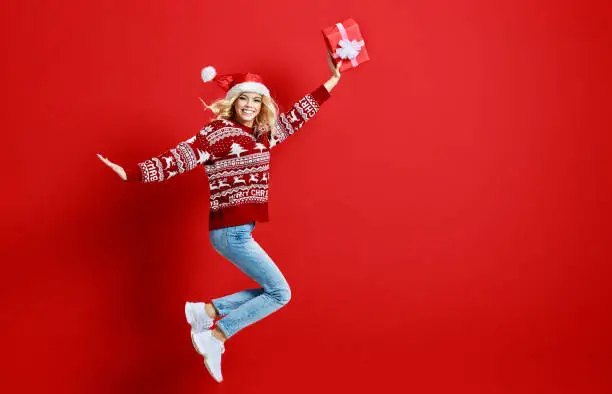 Photo of happy young cheerful girl laughs and jumps in christmas hat and with  gift on  red   background
