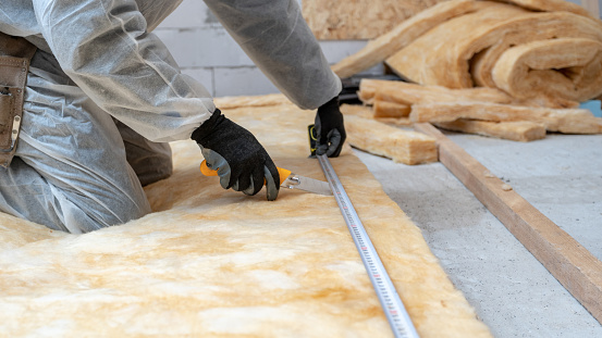 Cropped view of professional workman in protective workwear using knife and holding measuring tape over material, installing thermal insulation rock wool under the roof