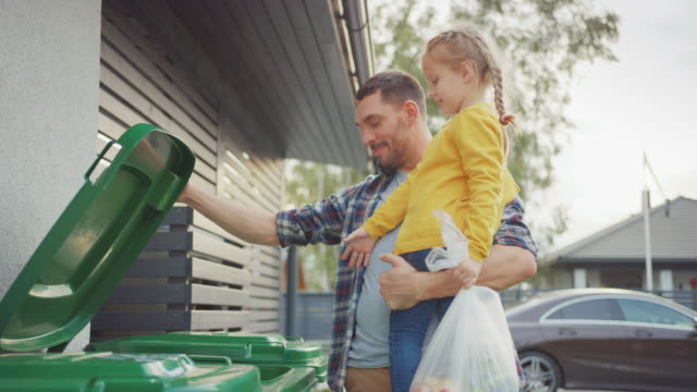 Happy Father Holding a Young Girl and Going to Throw Away an Empty Bottle and Food Waste into the Trash. They Use Correct Garbage Bins Because This Family is Sorting Waste and Helping the Environment.