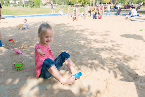 Little girl with a hand disability uses her feet to pick up a toy while playing in a sandbox.