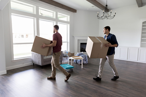Young male friends carry boxes into the living room of a new home.