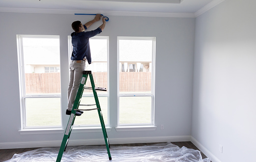 Young man places painter's tape on the crown moulding in a room in his new home. He is preparing the room for a fresh coat of paint. He is standing on a ladder in front of windows.