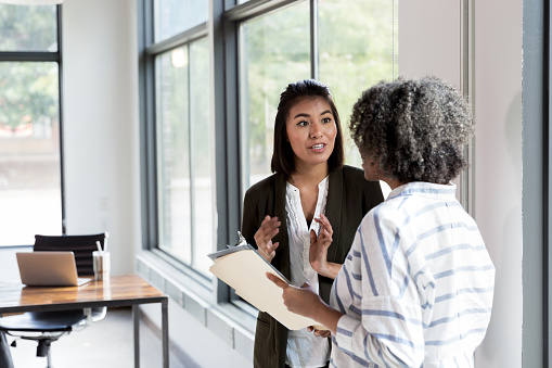 Mature adult female manager and mid adult female employee stand in the conference room and discuss staffing issues.