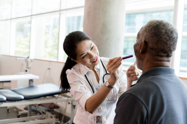 Female doctor examines a senior man's throat Female doctor uses a pen light and tongue depressor to examine a senior man's throat. emergency medicine stock pictures, royalty-free photos & images