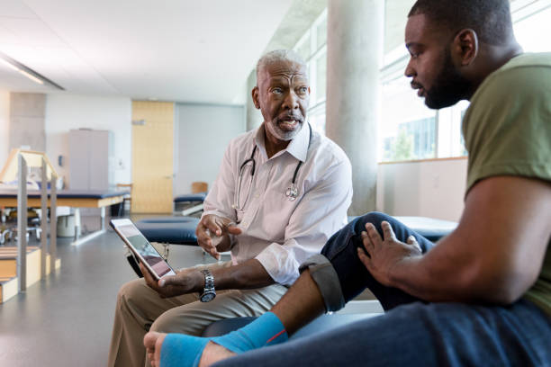 Male orthopedic doctor shows exercises to patient on digital tablet Serious male orthopedic doctor uses a digital tablet to show an injured male patient a video about appropriate exercises to strengthen the patient's food and ankle. The patient's food and ankle are wrapped with kinesiology tape. black military man stock pictures, royalty-free photos & images