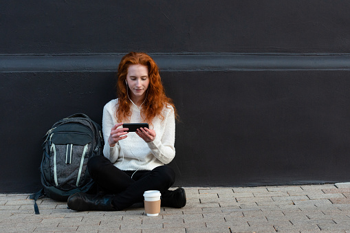 The young female student with a back pack and a cup of coffee sits on the ground and watches a video on her smart phone.