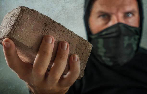 young man as fanatic and aggressive anarchist rioter . furious and scary violent anti-system protester in face mask throwing brick looking hostile at fighting riot in radical demonstration stock photo
