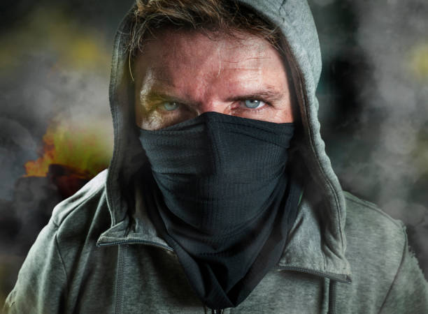 young man as ultra and radical anarchist rioter . furious antifa protester in face mask hostile on street on demonstration in violet riot and chaos between fire and smoke stock photo