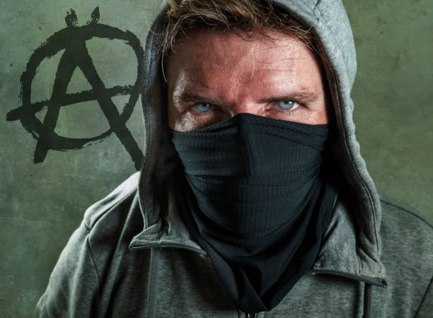young man as violent and ultra anarchist rioter . furious anti-system protester in face mask hostile at fighting riot in radical demonstration isolated on grey with anarchy symbol background stock photo