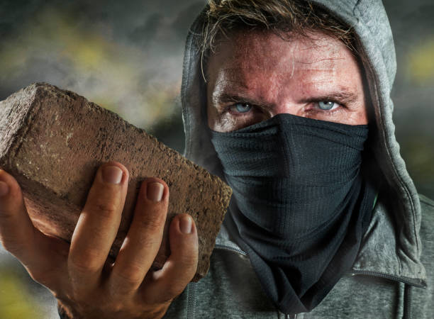 young man as ultra and radical anarchist rioter . furious antifa protester in face mask throwing brick hostile on street on demonstration in violet riot and chaos stock photo