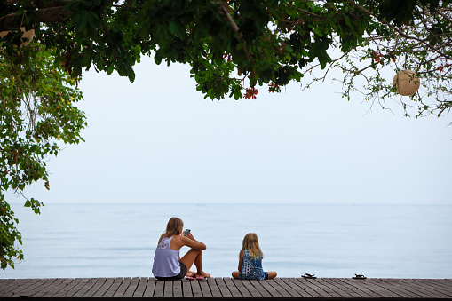 Happy mother, child relaxing and talking on luxury hotel veranda with beautiful view. People looking blue sea. Travel lifestyle, tropical beach leisure on family summer holiday with kids