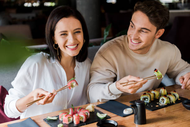 uomo felice e donna tenendo bacchette con sushi gustoso in ristorante - sushi chopsticks sushi bar food foto e immagini stock