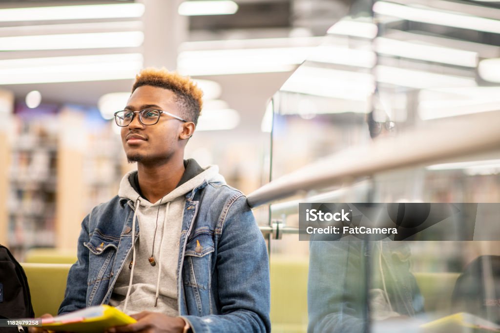 An African American University Student Studying in the Library stock photo An African American university student is sitting in the library with his textbooks open on his lap, and studying University Student Stock Photo