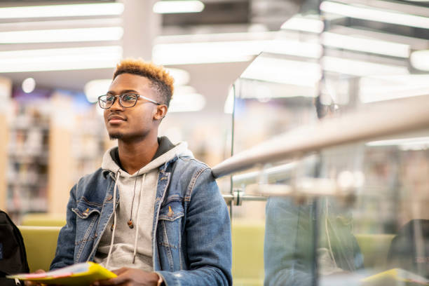 An African American University Student Studying in the Library stock photo An African American university student is sitting in the library with his textbooks open on his lap, and studying 20 29 years stock pictures, royalty-free photos & images