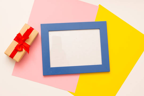 istock Christmas gift in craft paper with a red bow on a white background. Blue frame with space for your text. Pink and yellow color background. 1182480443