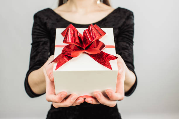 A girl in a black holiday dress holds a square white gift with a red bow. Girl gives a Christmas gift A girl in a black holiday dress holds a square white gift with a red bow. Girl gives a Christmas gift. gift lounge stock pictures, royalty-free photos & images