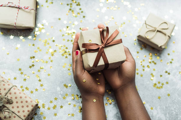 la ragazza nera fa un regalo fatto con le proprie mani. mani che tengono in mano un regalo artigianale con un nastro marrone su uno sfondo grigio con alcuni piccoli regali. laici piatti - holiday paper spotted close up foto e immagini stock