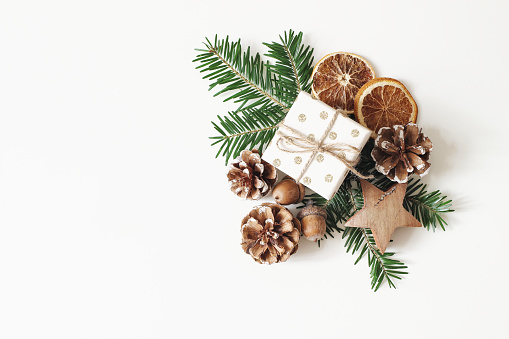 Christmas festive styled stock composition. Hand wrapped gift box, dry orange fruit slices, acorns, pine cones and fir tree branches isolated on white table background, winter flat lay, top view.