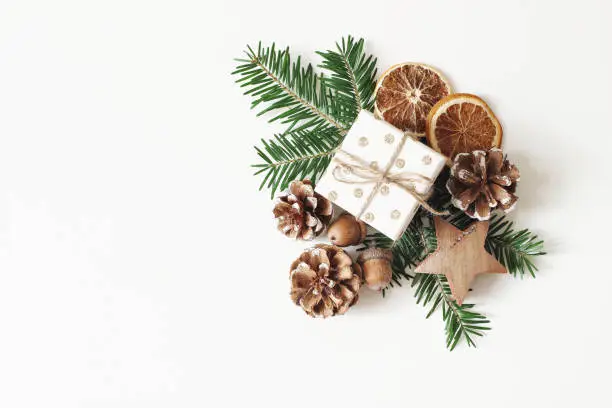 Photo of Christmas festive styled stock composition. Hand wrapped gift box, dry orange fruit slices, acorns, pine cones and fir tree branches isolated on white table background. Winter flat lay, top view.
