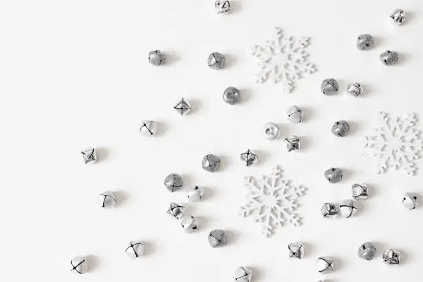 Photo of Christmas styled stock composition. Glittering jingle bells, silver dust and snowflakes decoration isolated on white table background. Flat lay, top view. Winter decorative pattern.