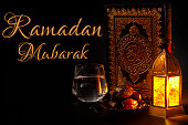 Ramadan Mubarak, muslim religious tradition, holy month of Islam and Iftar concept theme with a bowl of dates, prayer beads, glass of water, Quran and arabic lantern on black background