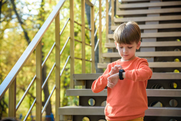 un ragazzo carino che indossa una camicia elegante sta vicino a scuola, guardando il suo orologio intelligente che tocca lo schermo. un bambino che usa un dispositivo elettronico. concetto di stile di vita - gps watch foto e immagini stock