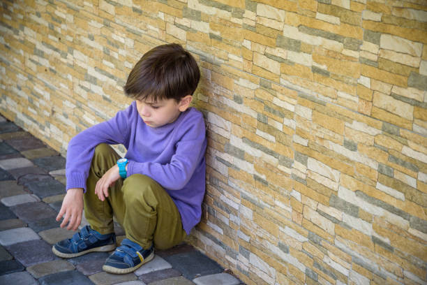 o menino pequeno triste que senta-se sozinho na escola esconde sua cara. conceito de isolamento e bullying. garoto triste e infeliz, criança estava chorando, chateado, se sentir doente - bullying child teasing little boys - fotografias e filmes do acervo