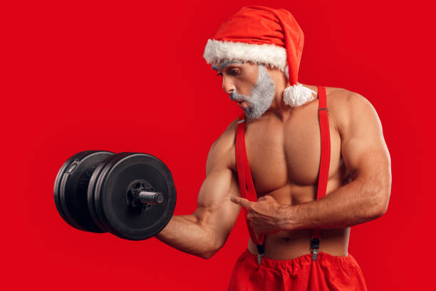 istock Attractive Santa Claus training with dumbbell, pointing at biceps 1182516120