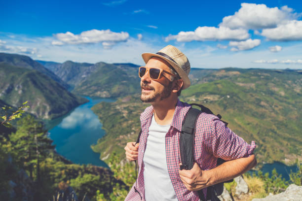 istock Fashionable backpacker on cliff against lake or river surrounded by hills. 1182525373