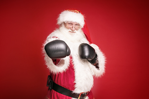 Real Santa Claus in boxing gloves is ready to fight , looking at camera, standing on red studio background. Merry Christmas and Happy New Year concept.