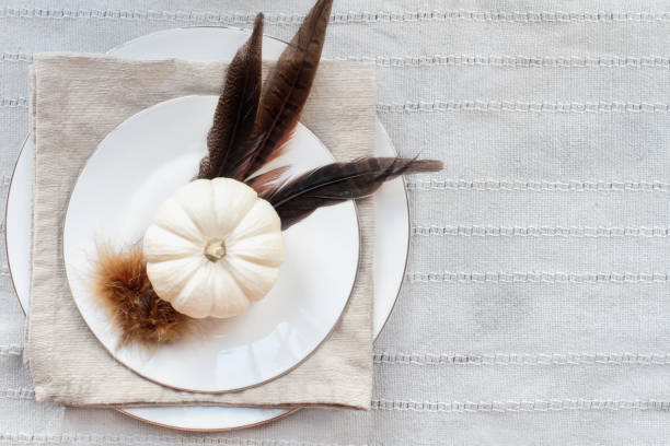 Thanksgiving Day place setting with white plates, mini white pumpkins, Pheasant feathers and napkin over grey table runner.