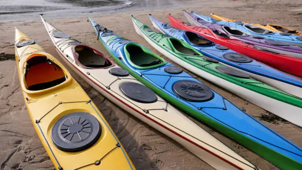 Brightly coloured carbon fibre sea kayaks lined up on a sandy beach, close to the water's edge. The vibrant colours of yellow, cream, blue, green, red and purple make a cheerful scene. The empty kayaks have been lined up ready for weekend activities, exploring the local coastline.